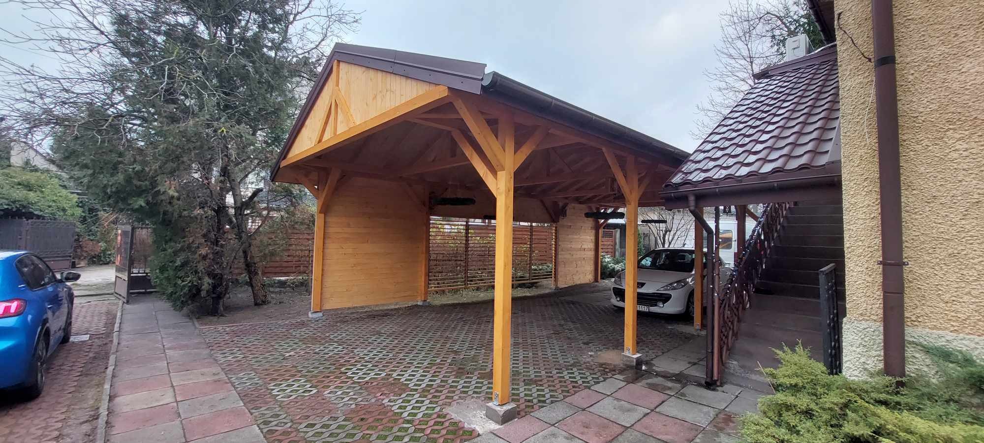 Drewniana konstrukcja zadaszenia dla dwóch samochodów, z brązowym dachem i podłogą z ażurowych płyt chodnikowych, widok z zewnątrz na tle domu i zieleni.