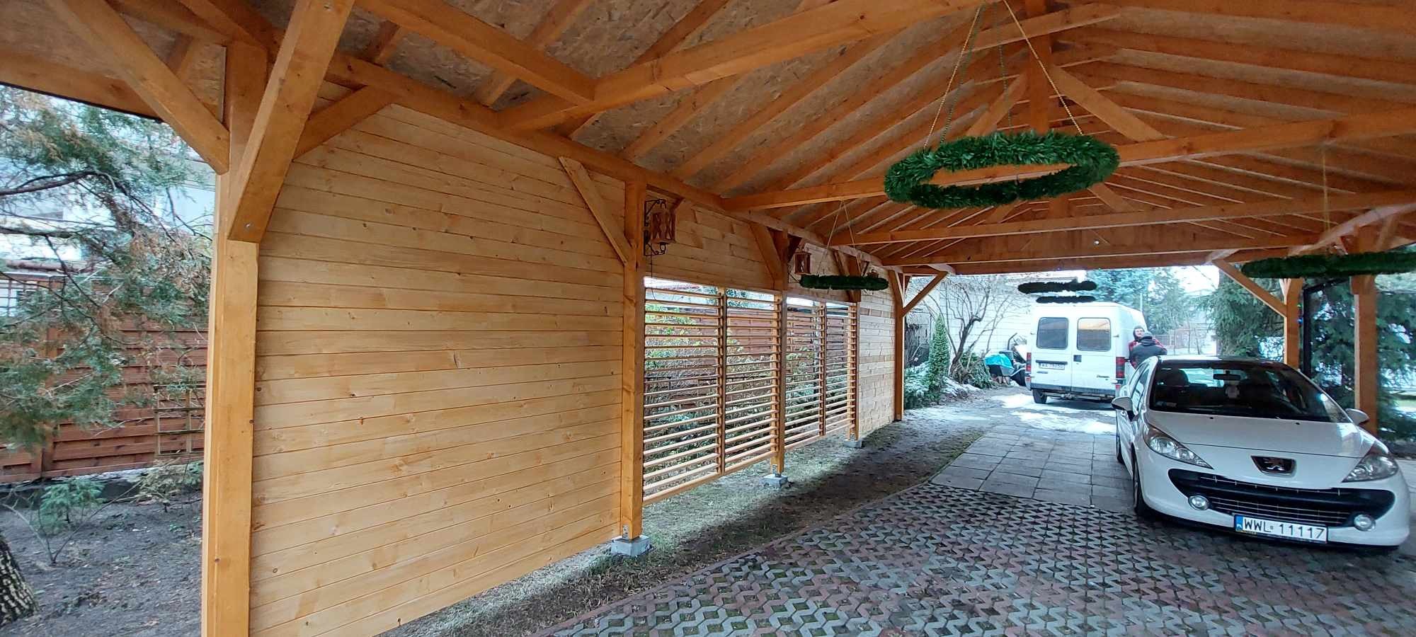 Zadaszone miejsce parkingowe z drewnianą konstrukcją, ścianami z desek i dekoracyjnymi zielonymi wieńcami pod sufitem, na zewnątrz zaparkowany biały samochód.