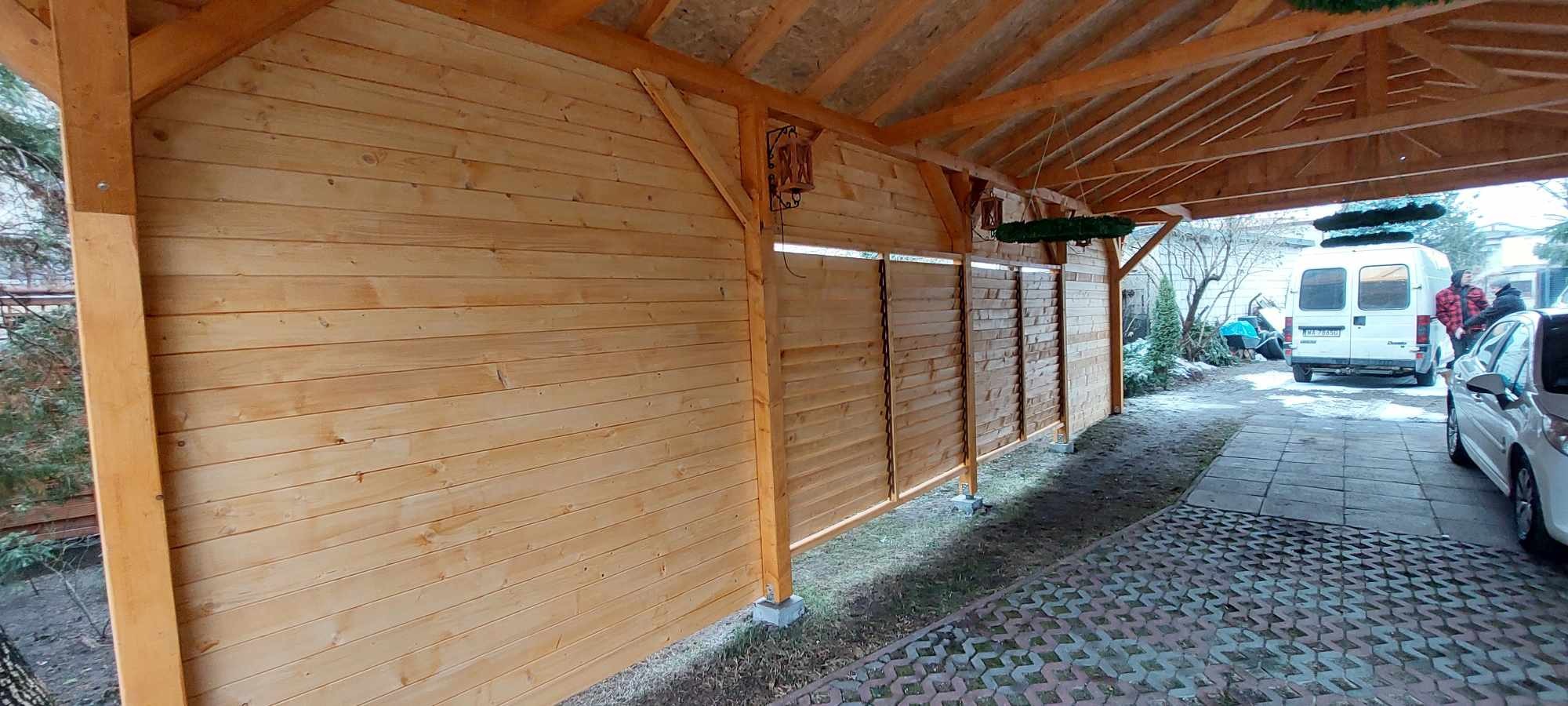 Zadaszenie tarasu z drewnianą ścianą działową z poziomych desek, oświetlone lampami w stylu retro, widoczne samochody na posesji.