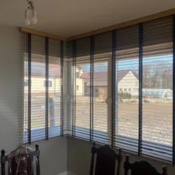 Drewniane żaluzje poziome w oknie narożnym, widok na dom z ogrodem w słoneczny dzień.