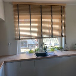 Wiktor Kurłowicz - Drewniane lamele w oknie kuchennym, czarne linki podtrzymujące, widok na zewnątrz z budową w tle, zlewozmywak z kwiatami na parapecie.