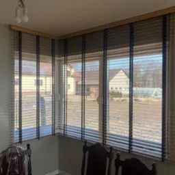 Drewniane żaluzje poziome w narożnym oknie z widokiem na domy i ogród z tunelem foliowym.