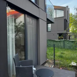 Zewnętrzna moskitiera na oknie tarasowym nowoczesnego domu w szarej tonacji. Widok z zewnątrz na ciemne meble balkonowe i zielony ogród.