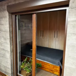 Brązowa roleta zewnętrzna częściowo zasłonięta, widok z zewnątrz na pokój z łóżkiem i szafą. Roślina w doniczce na pierwszym planie, drewniane wykończenie.