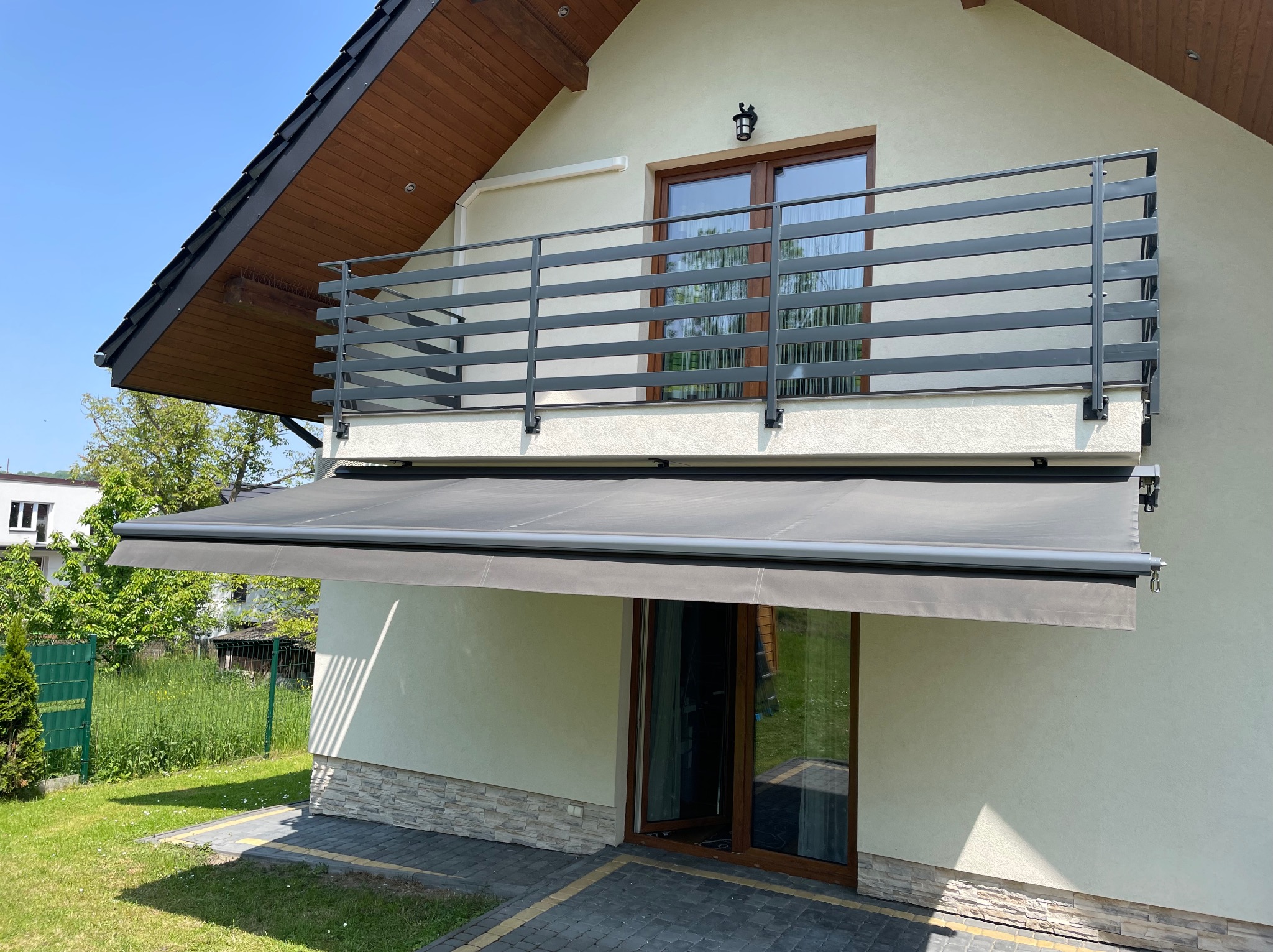 Szara markiza tarasowa zamontowana nad przeszklonymi drzwiami balkonowymi w nowoczesnym domu z szarą balustradą balkonową.