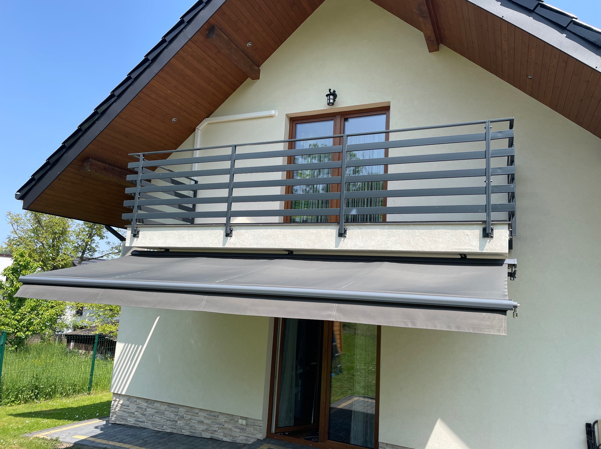 Szara markiza zamontowana nad drzwiami balkonowymi w nowoczesnym domu z drewnianą podbitką dachu i metalową balustradą balkonową.