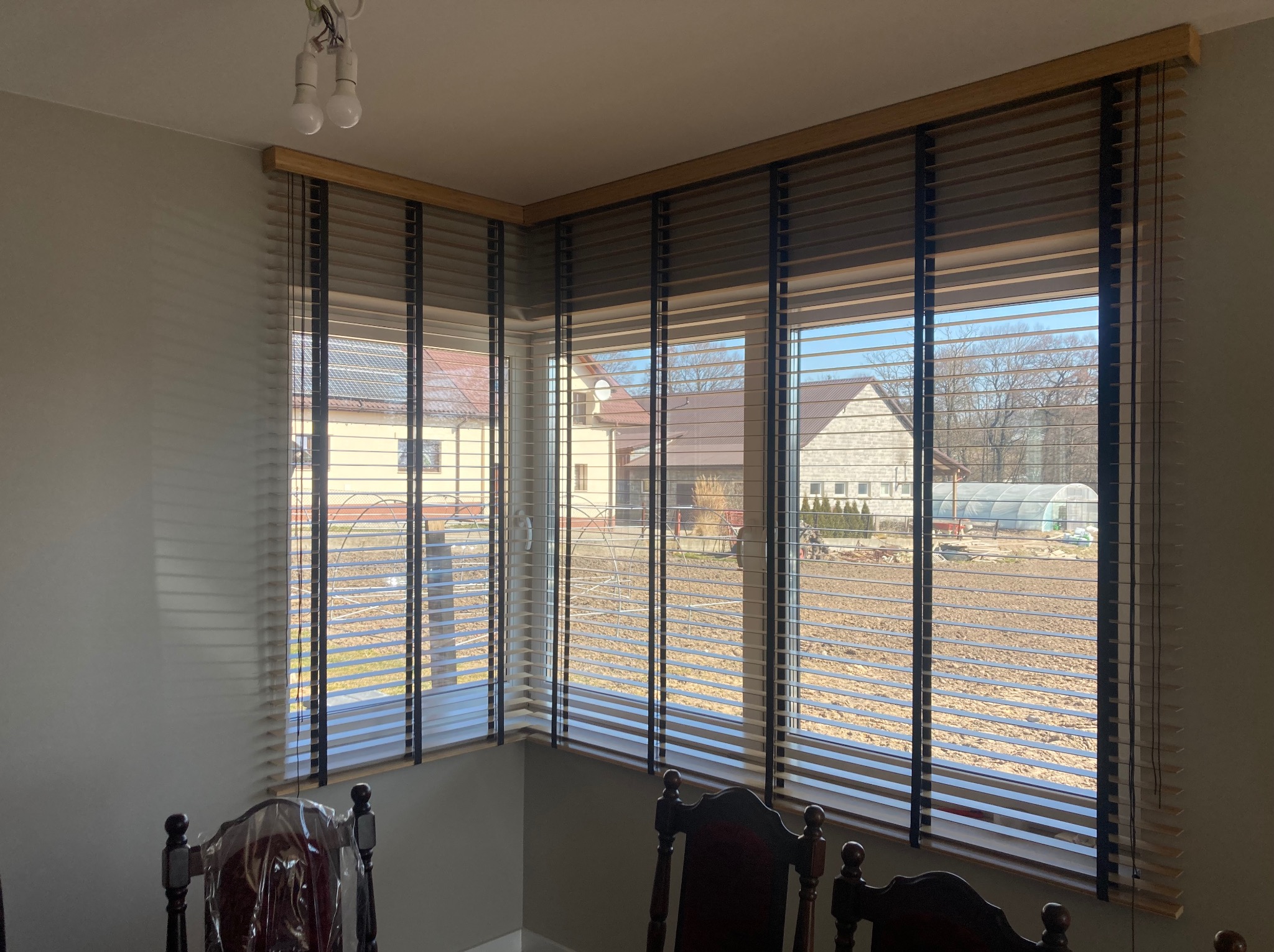 Drewniane żaluzje poziome w oknie narożnym, widok na dom z ogrodem w słoneczny dzień.