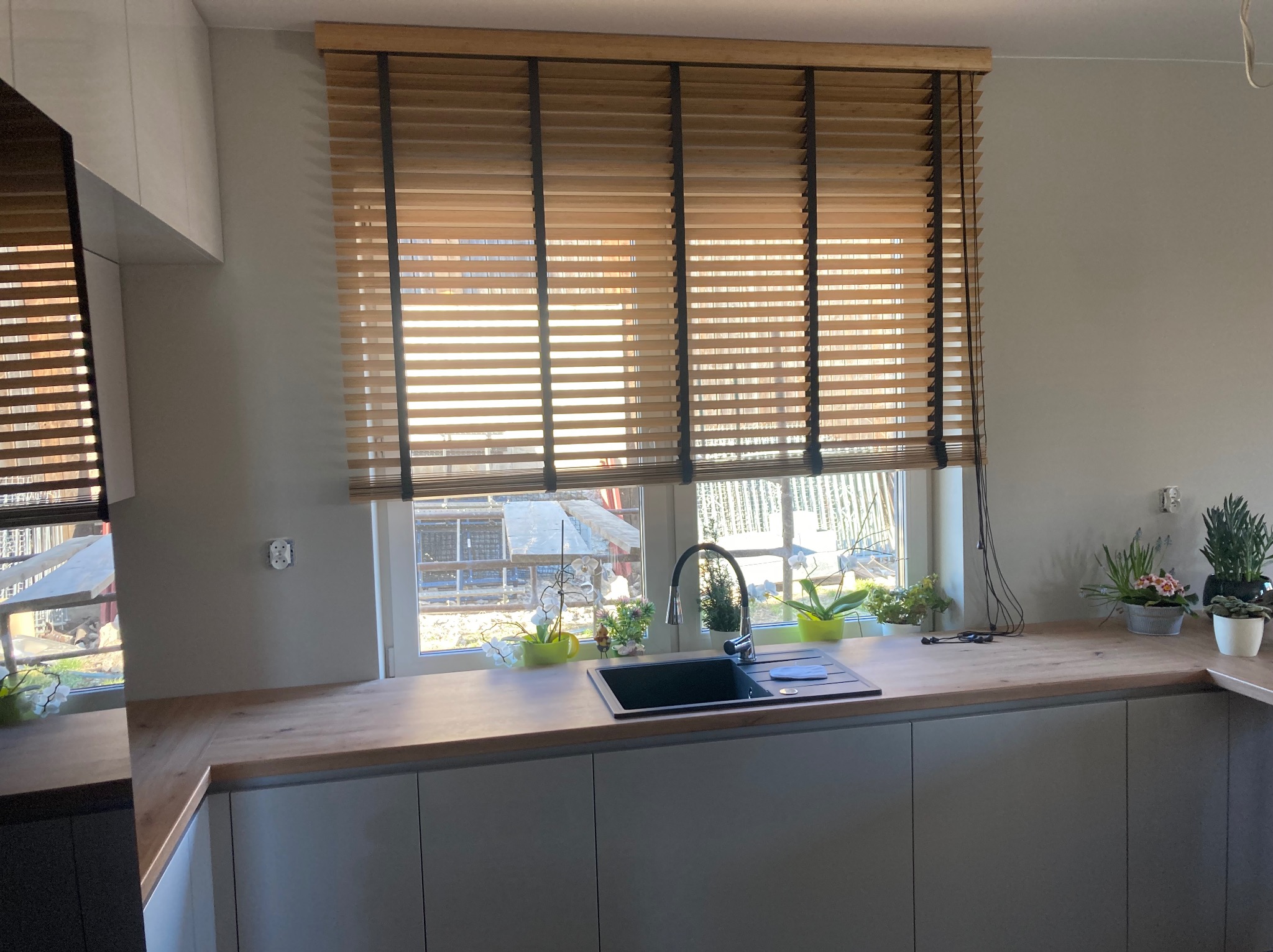 Drewniane lamele w oknie kuchennym, czarne linki podtrzymujące, widok na zewnątrz z budową w tle, zlewozmywak z kwiatami na parapecie.