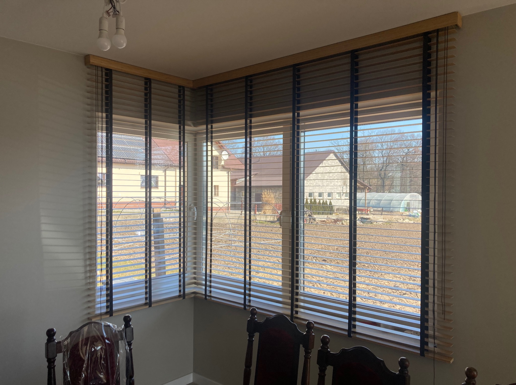 Drewniane żaluzje poziome w narożnym oknie z widokiem na domy i ogród z tunelem foliowym.