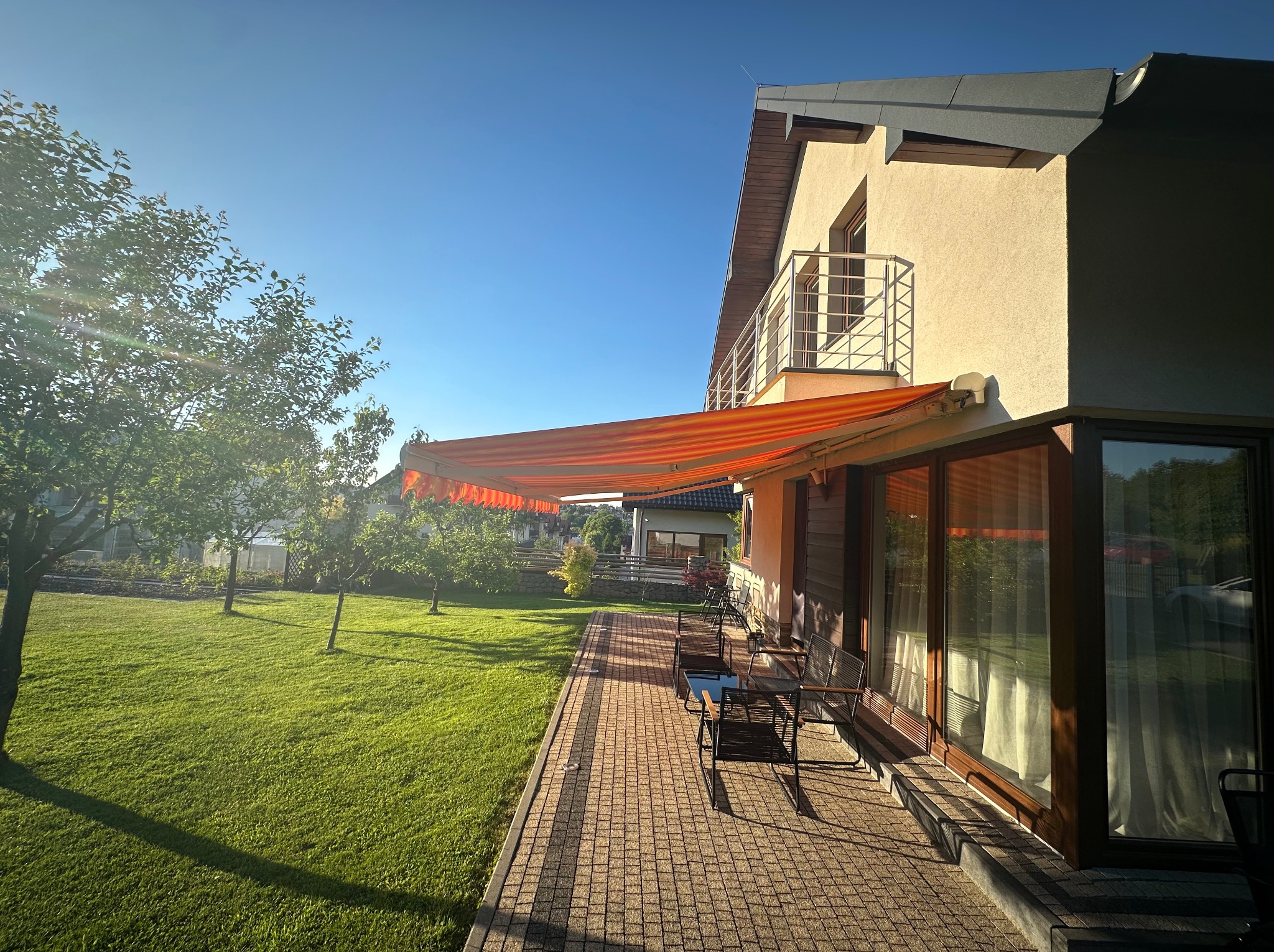 Pomarańczowa markiza tarasowa rozciągnięta nad patio z meblami ogrodowymi, widok na zadbany trawnik i dom w słoneczny dzień.