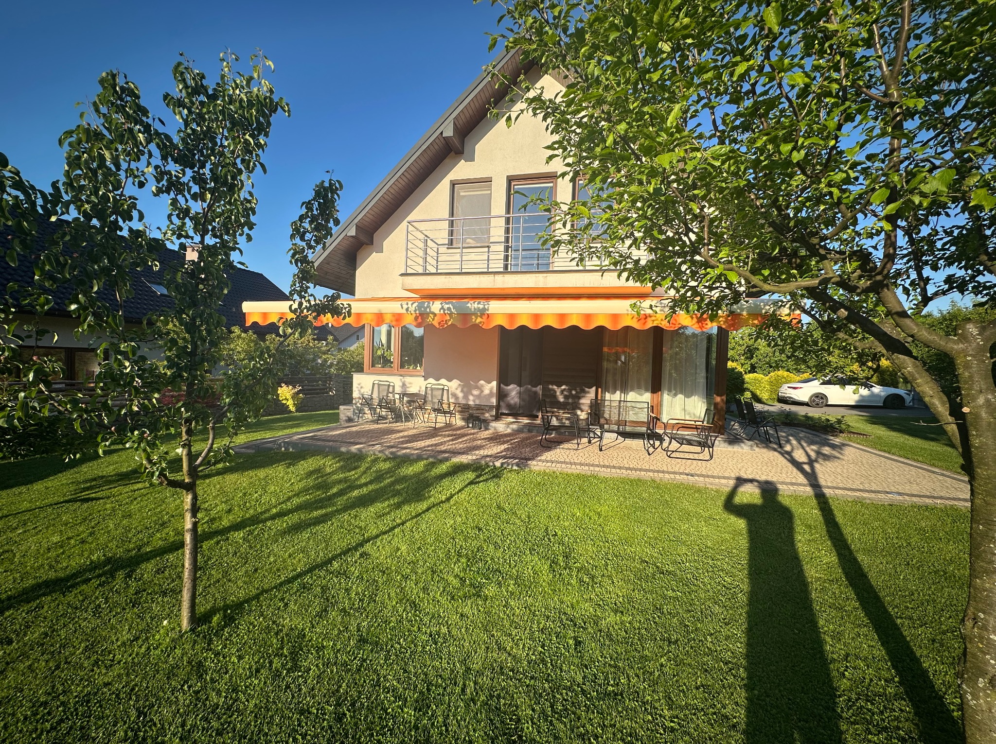 Pomarańczowa markiza nad tarasem domu w słoneczny dzień. Na pierwszym planie cień osoby robiącej zdjęcie. W tle widoczny biały samochód.