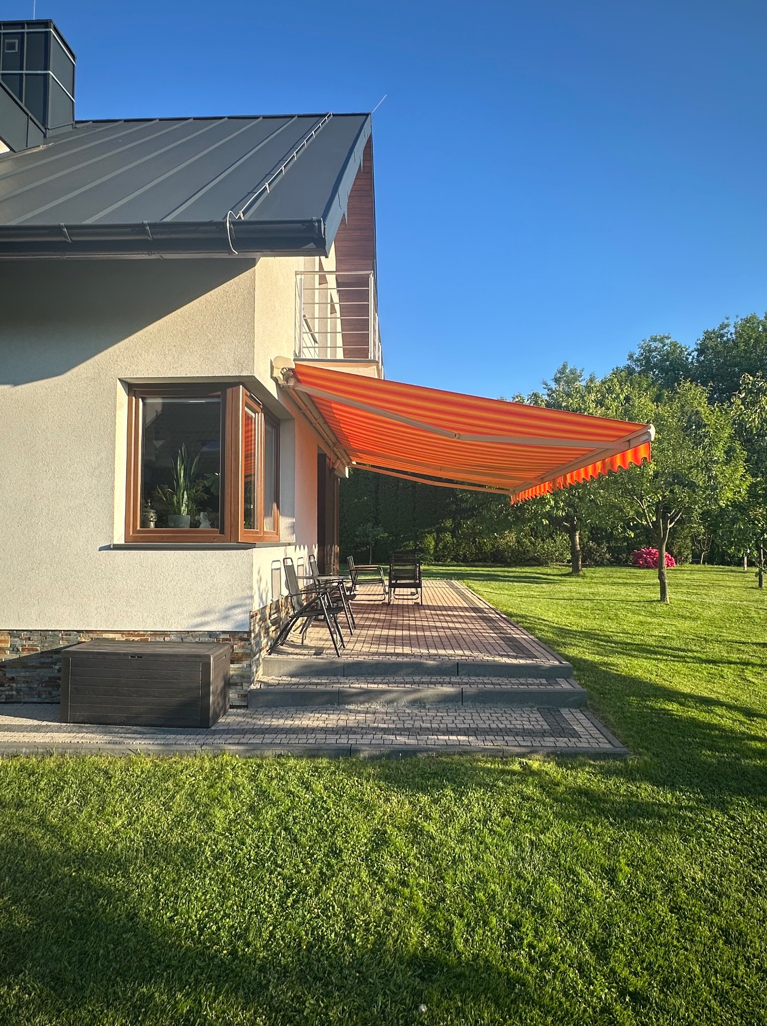 Rozłożona markiza w paski, chroniąca taras domu z zadbanym trawnikiem. Widoczne meble ogrodowe i okno z roślinami. Słoneczny dzień.