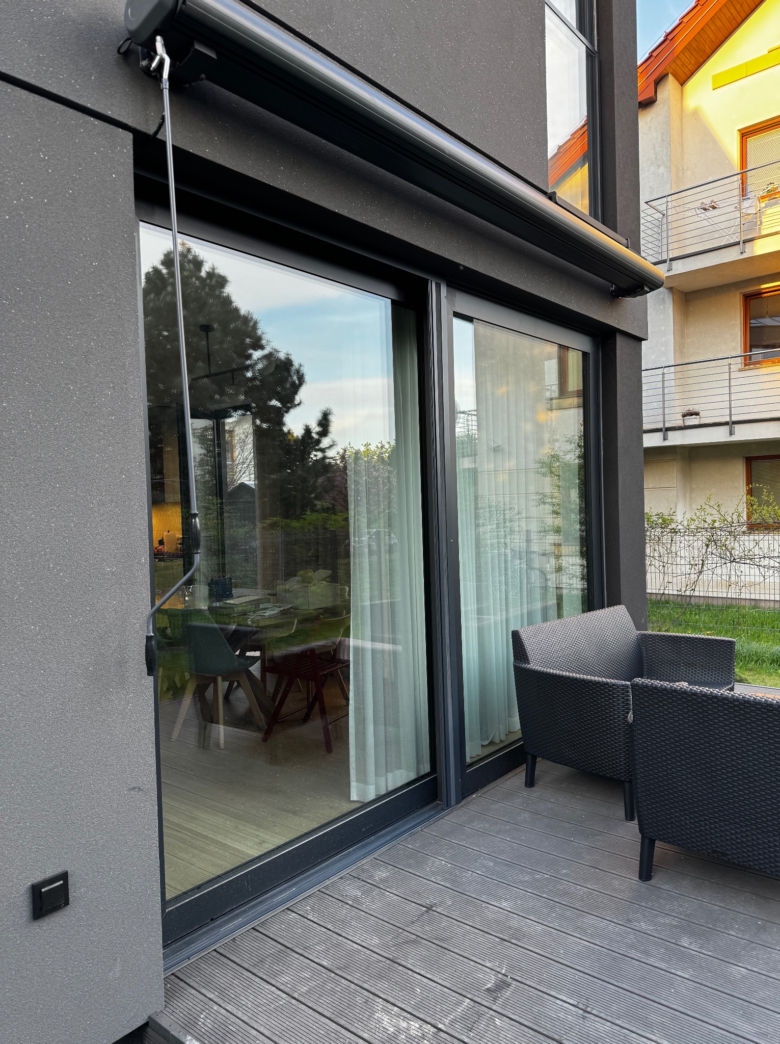 Elegancka, czarna markiza tarasowa zamontowana nad dużym, przesuwnym oknem. Widoczne czarne meble ogrodowe i drewniany taras na tle nowoczesnego budynku.