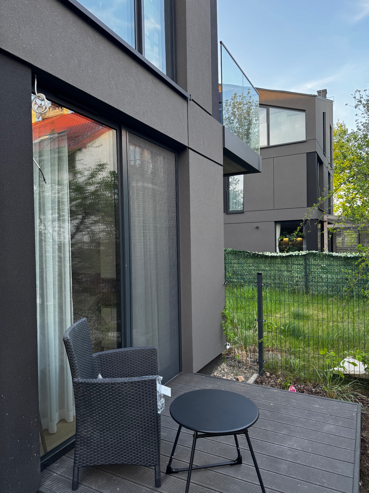 Zewnętrzna moskitiera na oknie tarasowym nowoczesnego domu w szarej tonacji. Widok z zewnątrz na ciemne meble balkonowe i zielony ogród.