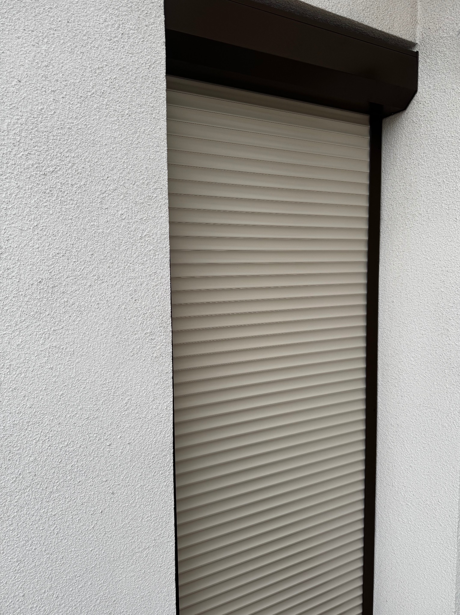 Zamknięta roleta zewnętrzna, beżowa, z ciemnobrązową ramą, na tle białego, chropowatego muru. Widok z dołu, ukazujący szczegóły konstrukcji i fakturę.