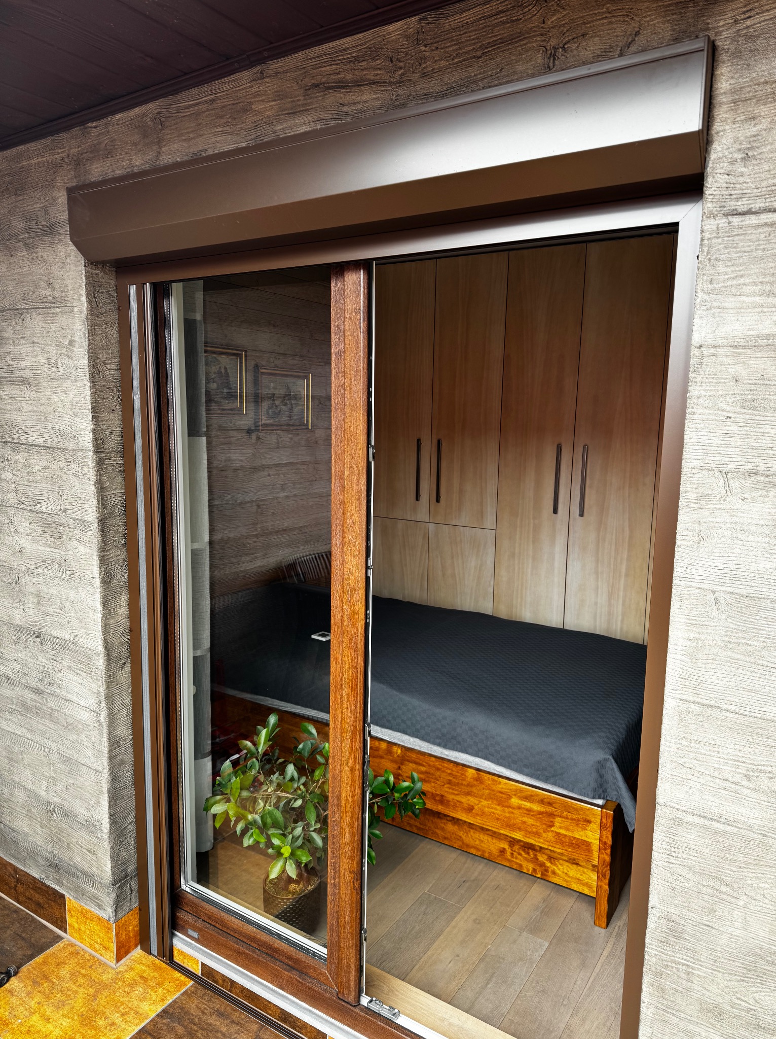 Brązowa roleta zewnętrzna częściowo zasłonięta, widok z zewnątrz na pokój z łóżkiem i szafą. Roślina w doniczce na pierwszym planie, drewniane wykończenie.