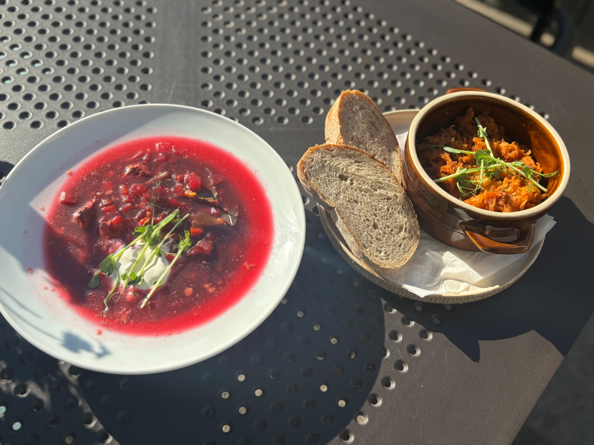 Bordosz w białym talerzu z kleksem śmietany i kiełkami, bigos w brązowym garnku i trzy kromki chleba na tle ażurowego stolika.