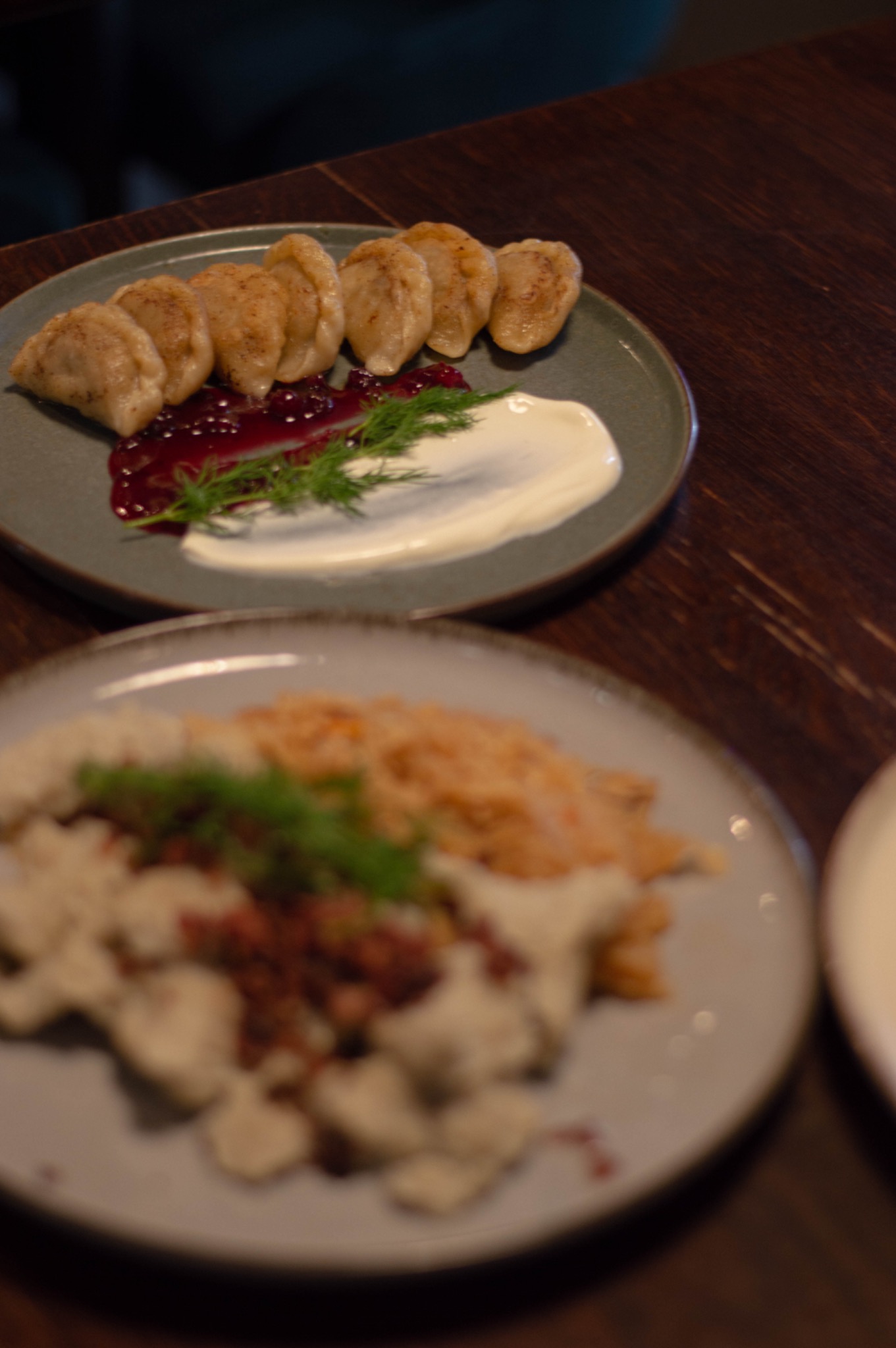 Dwa talerze z pierogami na drewnianym stole: na pierwszym talerzu pierogi z owocami i śmietaną, udekorowane koperkiem, na drugim talerzu pierogi z kapustą i skwarkami, również udekorowane koperkiem.