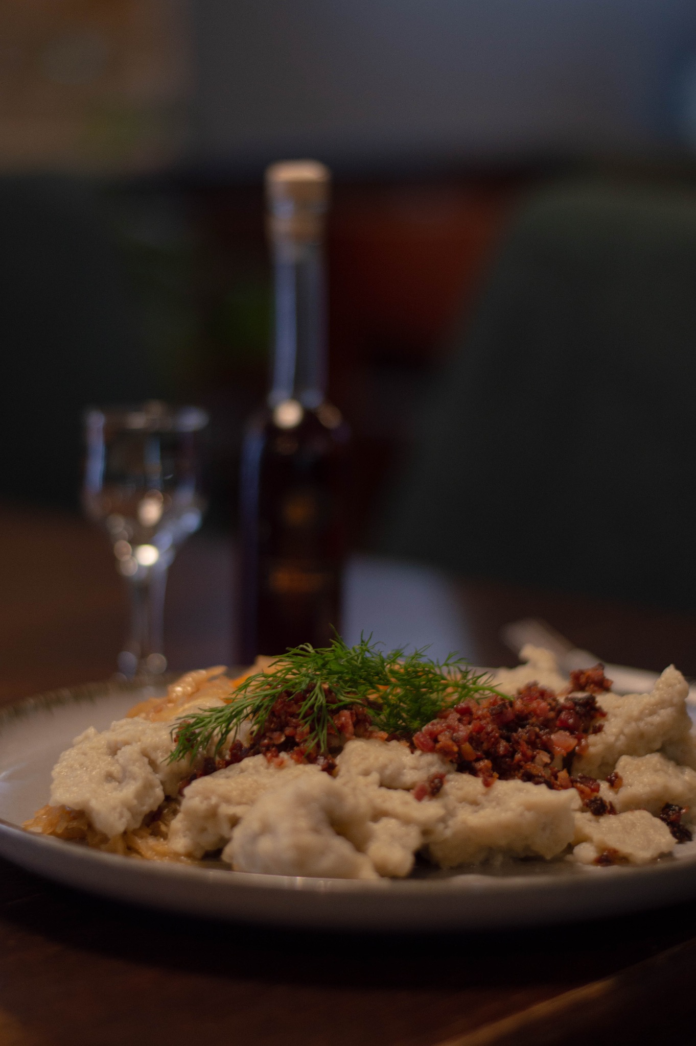 Kluski kładzione na kapuście, posypane skwarkami i udekorowane świeżym koperkiem, podane na talerzu; w tle szklanka i butelka, całość w stonowanej kolorystyce.