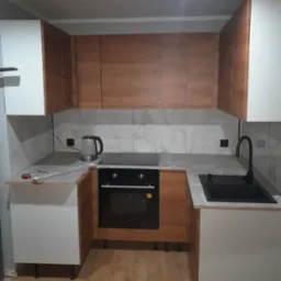 Nowoczesna kuchnia w trakcie montażu: biało-drewniane szafki, szary blat, czarny zlew i płyta indukcyjna, widoczne narzędzia na blacie, czajnik elektryczny.