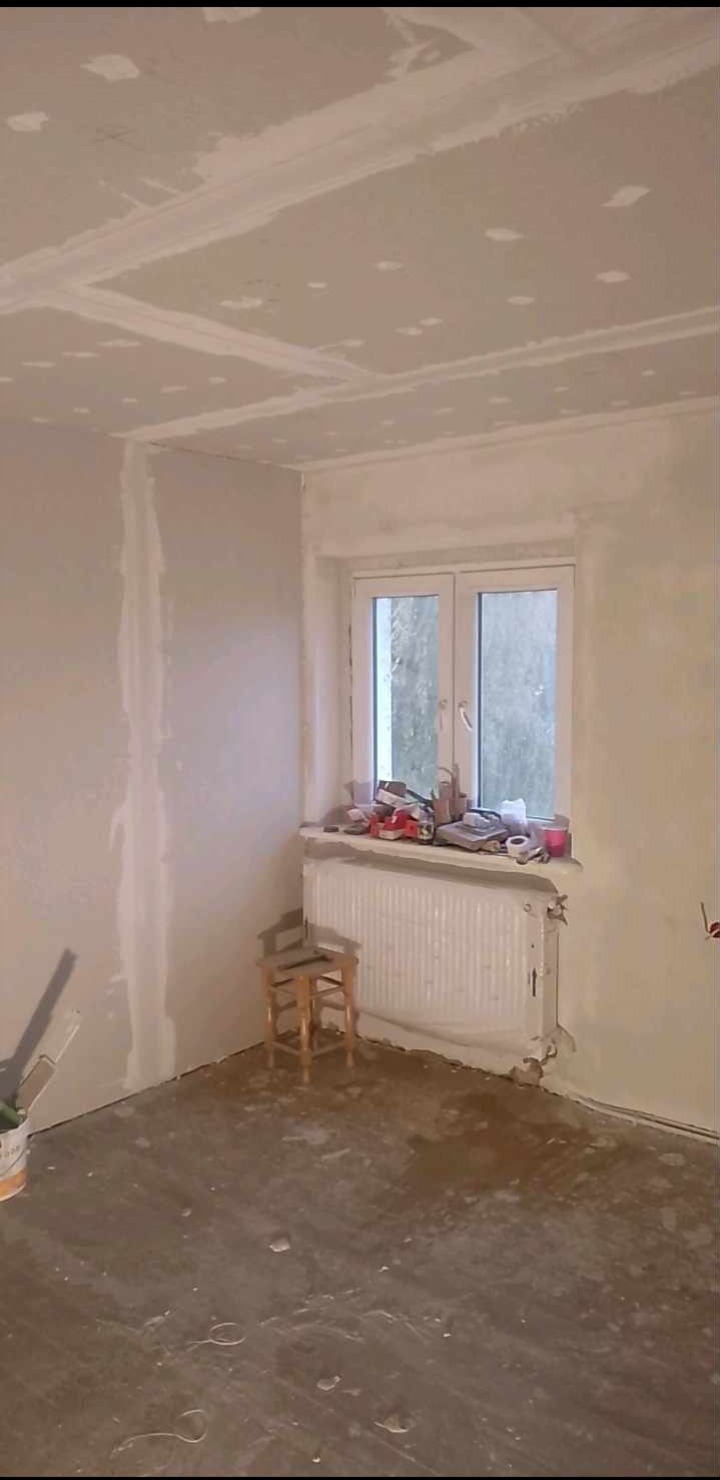 Pokój w trakcie prac wykończeniowych: ściany i sufit po szpachlowaniu, widoczny grzejnik, okno i mały stołek.