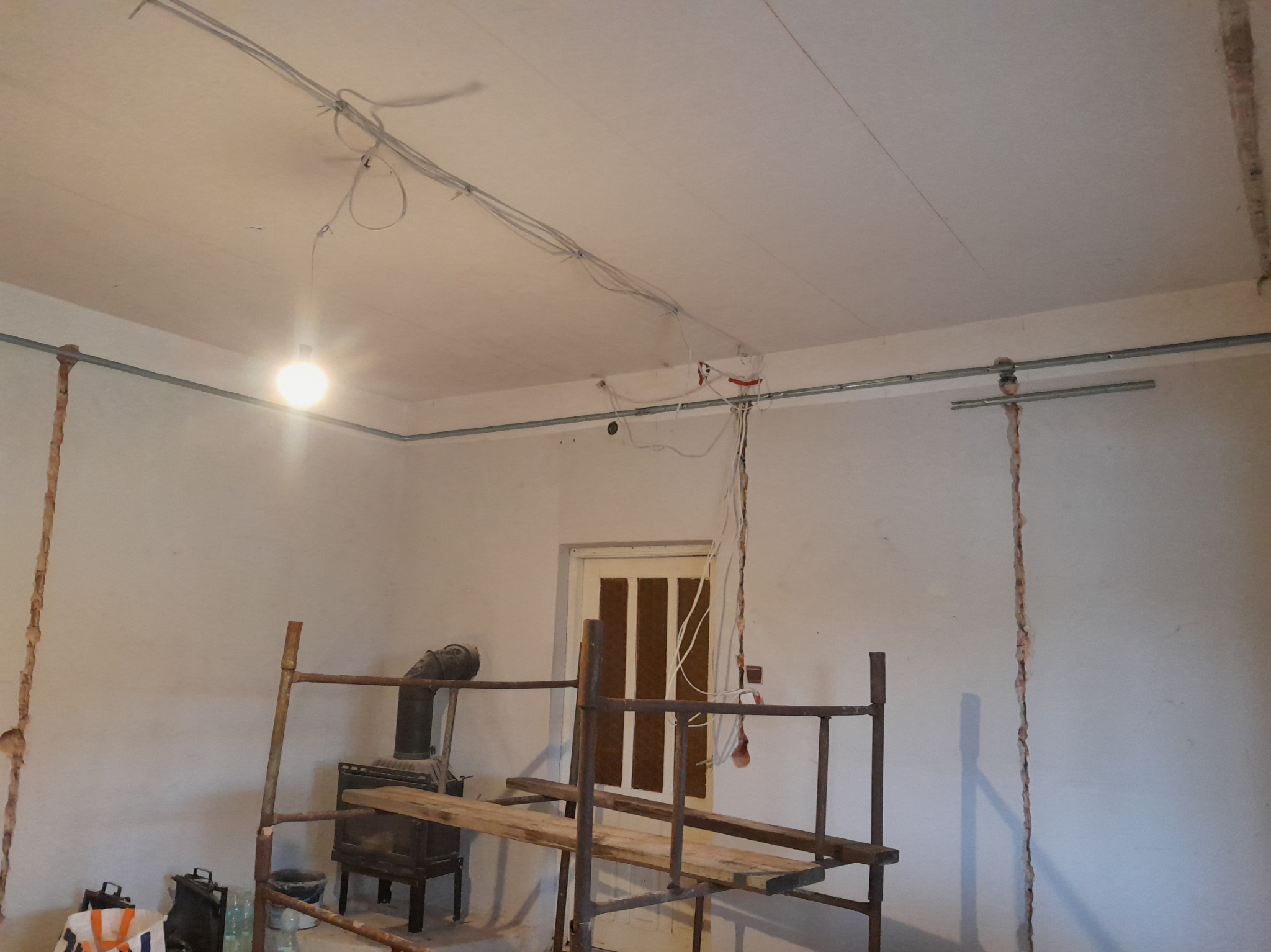 Wnętrze remontowanego pokoju z widocznymi przewodami elektrycznymi na suficie, rusztowaniem, piecem kaflowym i częściowo odspojoną warstwą tynku na ścianach.