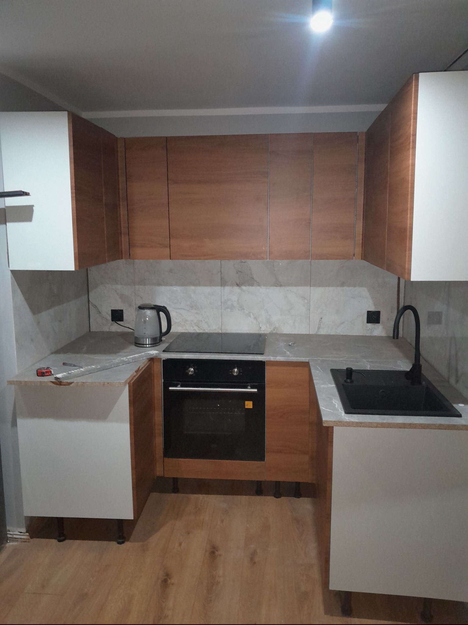 Nowoczesna kuchnia w trakcie montażu: biało-drewniane szafki, szary blat, czarny zlew i płyta indukcyjna, widoczne narzędzia na blacie, czajnik elektryczny.