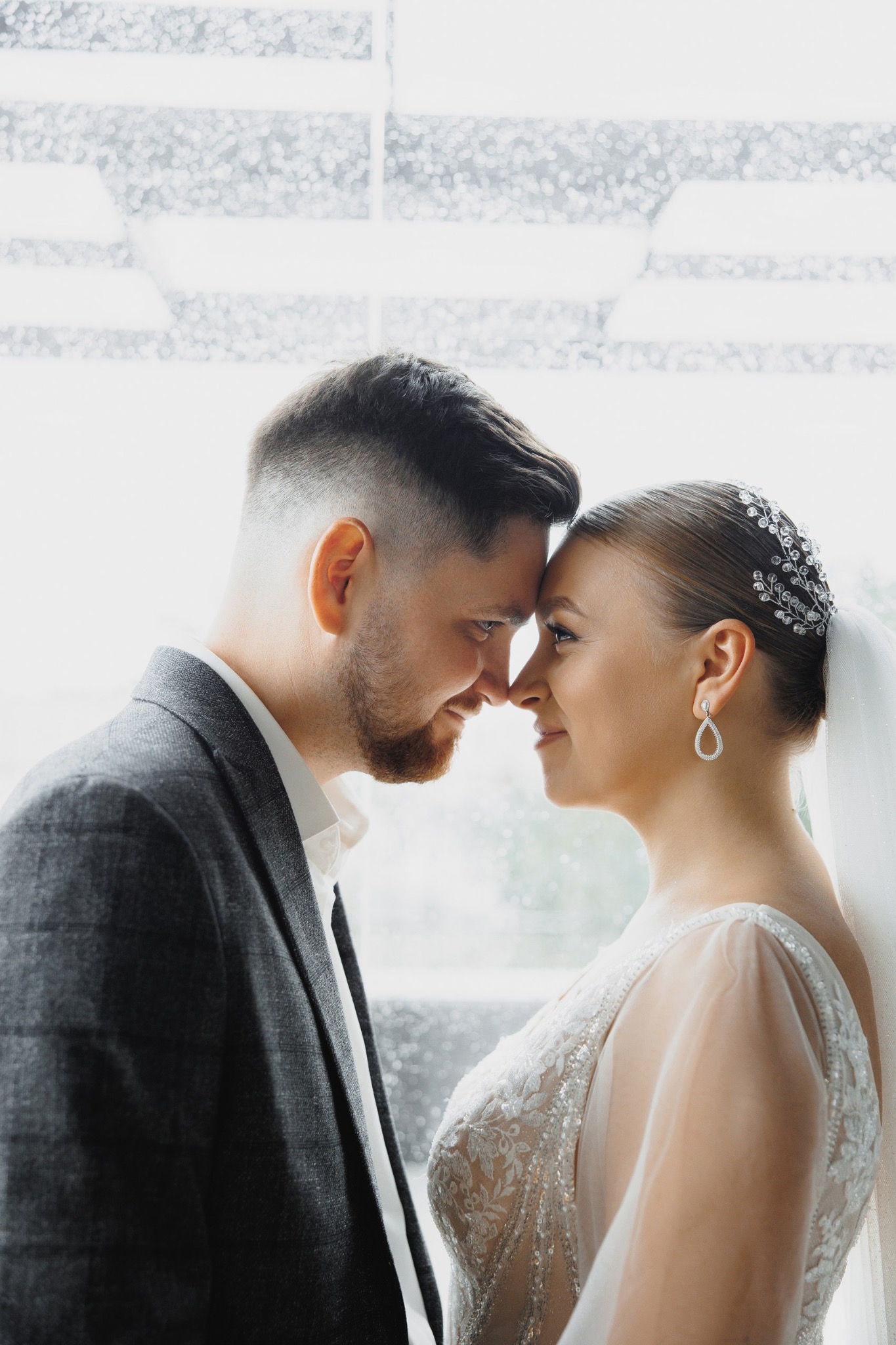 Młoda para, zbliżone profile, dotykają się nosami, ona w koronkowej sukni ślubnej z tiulowymi rękawami i ozdobą we włosach, on w szarym tweedowym garniturze, rozmyte tło okna.