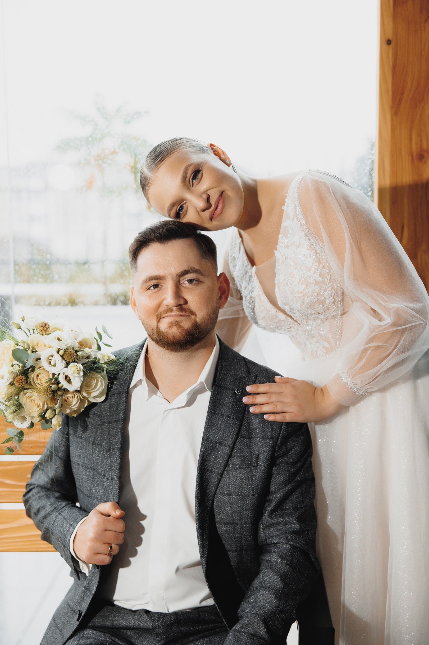 Para młoda: uśmiechnięta panna młoda opiera głowę na ramieniu pana młodego w szarym garniturze, bukiet białych róż, okno z kroplami deszczu w tle.