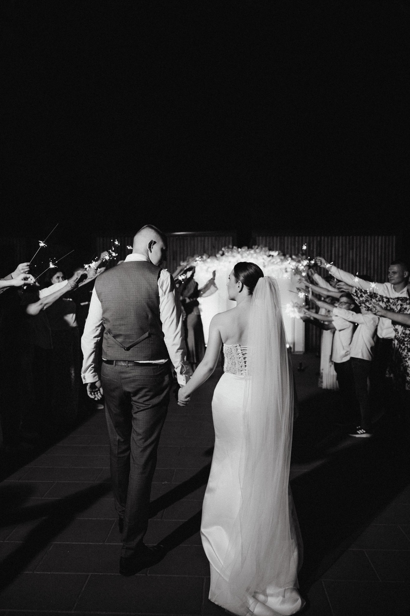 Para młoda trzymająca się za ręce, idąca pod szpalerem iskrzących się zimnych ogni, widziana od tyłu, w czerni i bieli.