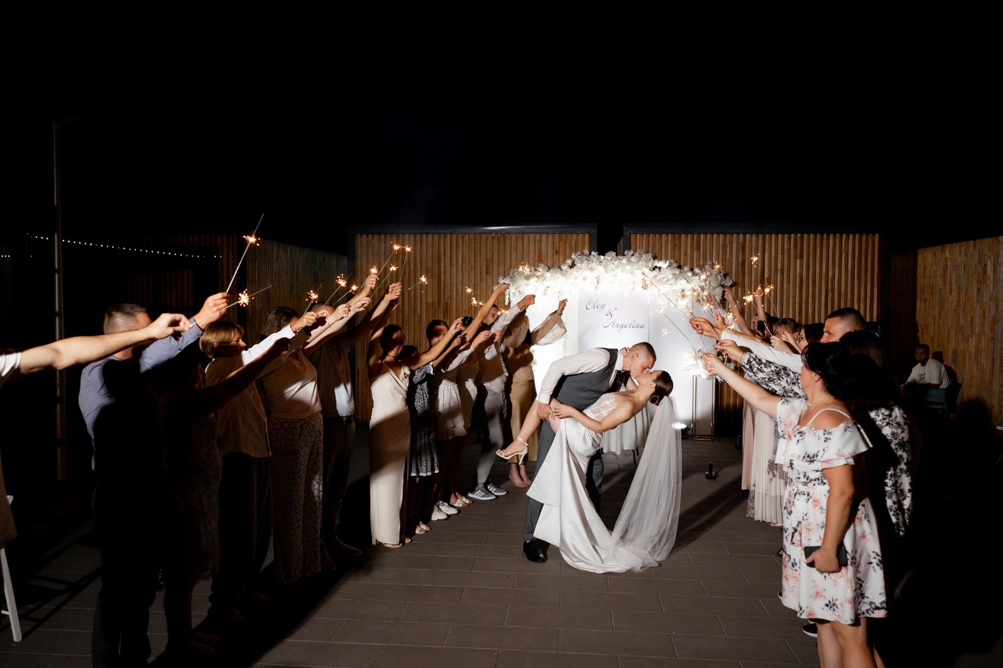 Para młoda w objęciach, unoszona przez pana młodego, otoczona gośćmi z zimnymi ogniami na tle dekoracji ślubnej z kwiatów i napisu Oleg & Angelina.