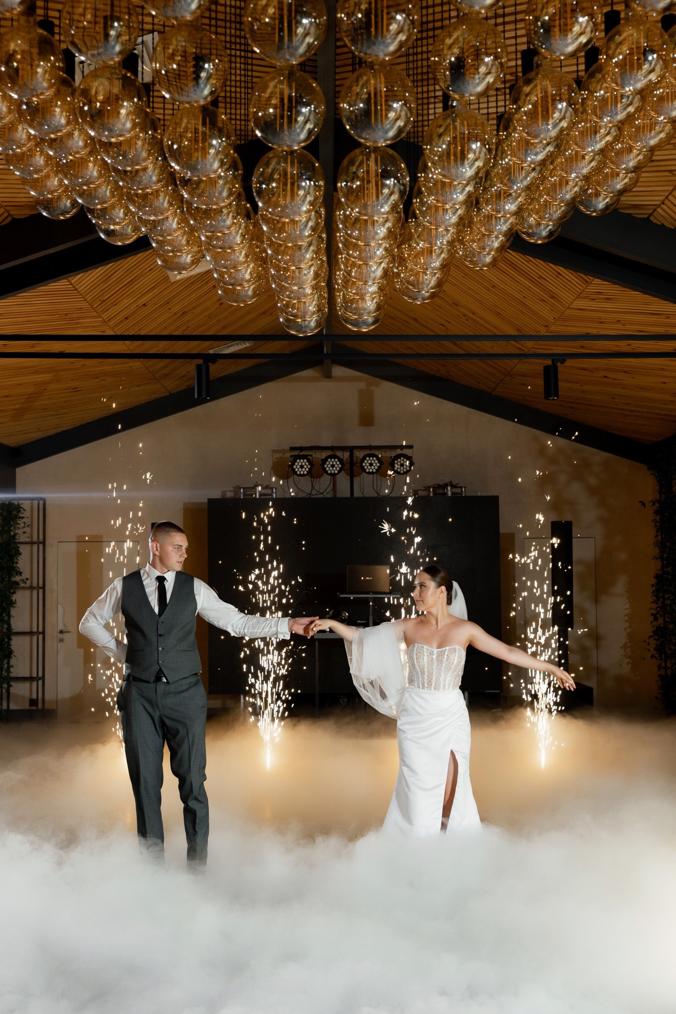Para młoda tańczy w dymie i blasku iskier podczas wesela, dekoracyjne oświetlenie sufitowe w tle.