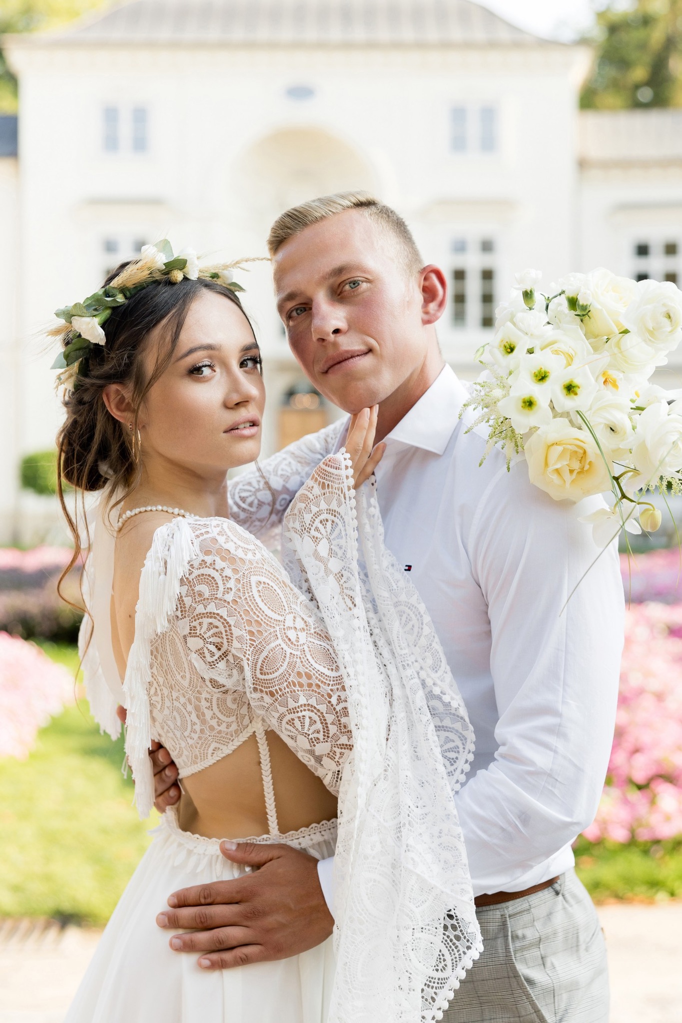 Młoda para w objęciach na tle jasnego budynku; panna młoda w koronkowej sukni z kwiatowym wiankiem, pan młody w białej koszuli trzymający bukiet białych kwiatów.