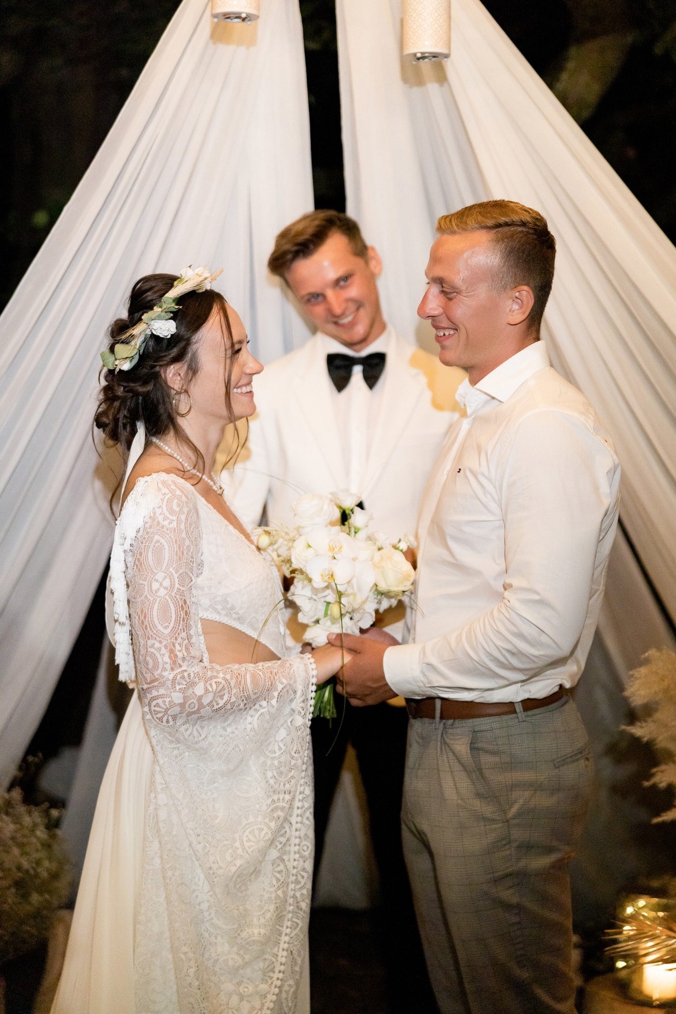 Uśmiechnięta para młoda, panna młoda w koronkowej sukni z kwiatowym wiankiem, pan młody w jasnej koszuli i kraciastych spodniach, za nimi mężczyzna w białym smokingu z czarną muchą, ceremonia...