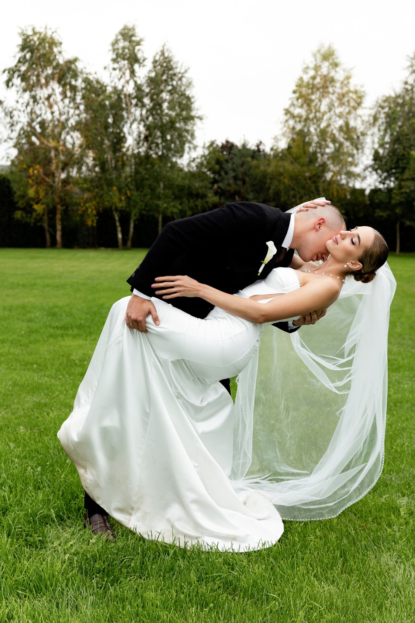 Panna młoda w białej sukni i welonie, unoszona przez pana młodego w czarnym smokingu, pocałunek na tle zielonego trawnika i drzew