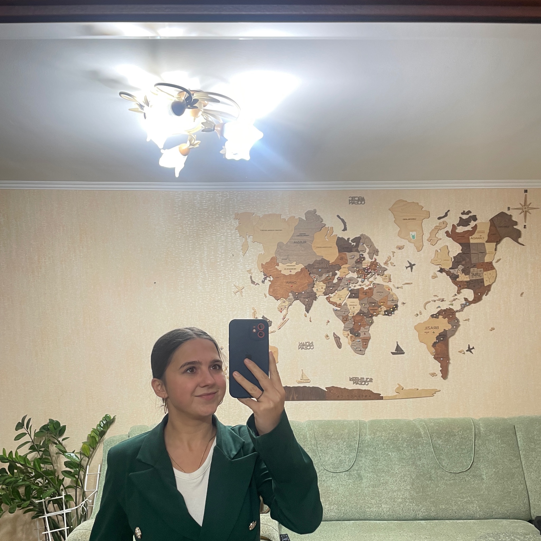 Kobieta w zielonej marynarce robi selfie na tle drewnianej mapy świata na ścianie, widoczny zielony narożnik sofy i roślina doniczkowa.
