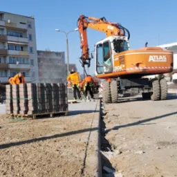 Pomarańczowy koparko-ładowarka Atlas 1304 na budowie, paleta z kostką brukową, robotnicy w pomarańczowych ubraniach, teren przygotowany pod ułożenie nawierzchni.