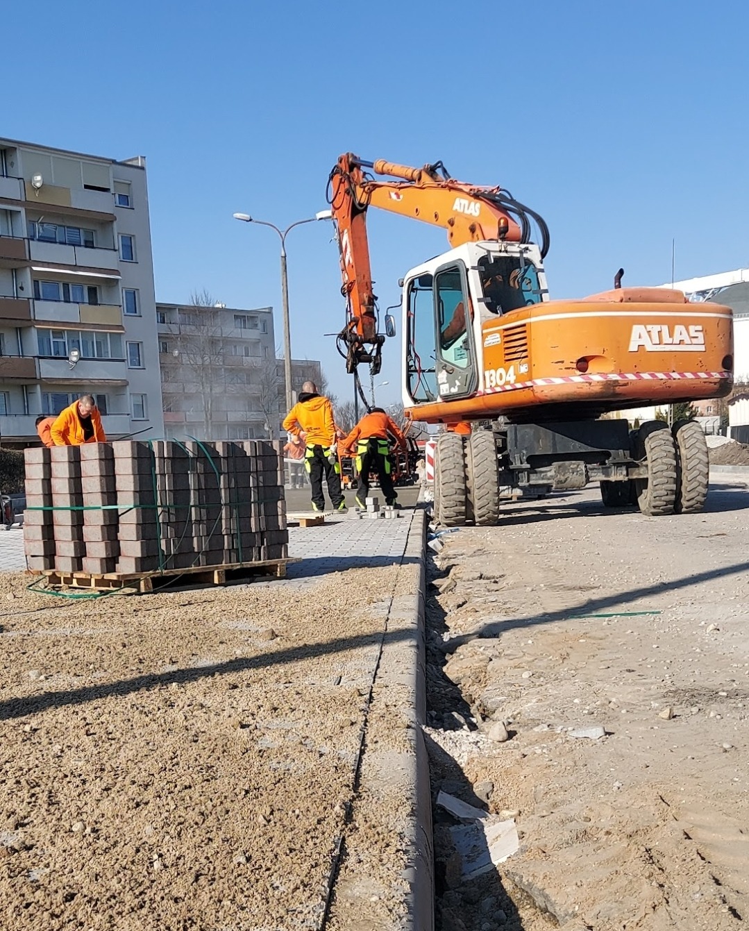 Pomarańczowy koparko-ładowarka Atlas 1304 na budowie, paleta z kostką brukową, robotnicy w pomarańczowych ubraniach, teren przygotowany pod ułożenie nawierzchni.