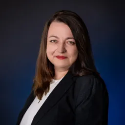 Portret kobiety w czarnej marynarce i białej bluzce, z delikatnym uśmiechem, na ciemnoniebieskim tle z subtelnym gradientem.