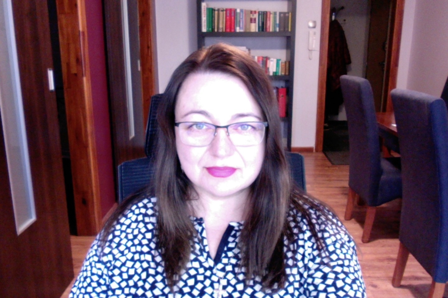 Portret kobiety w okularach, siedzącej na krześle biurowym, z biblioteczką i drzwiami w tle.