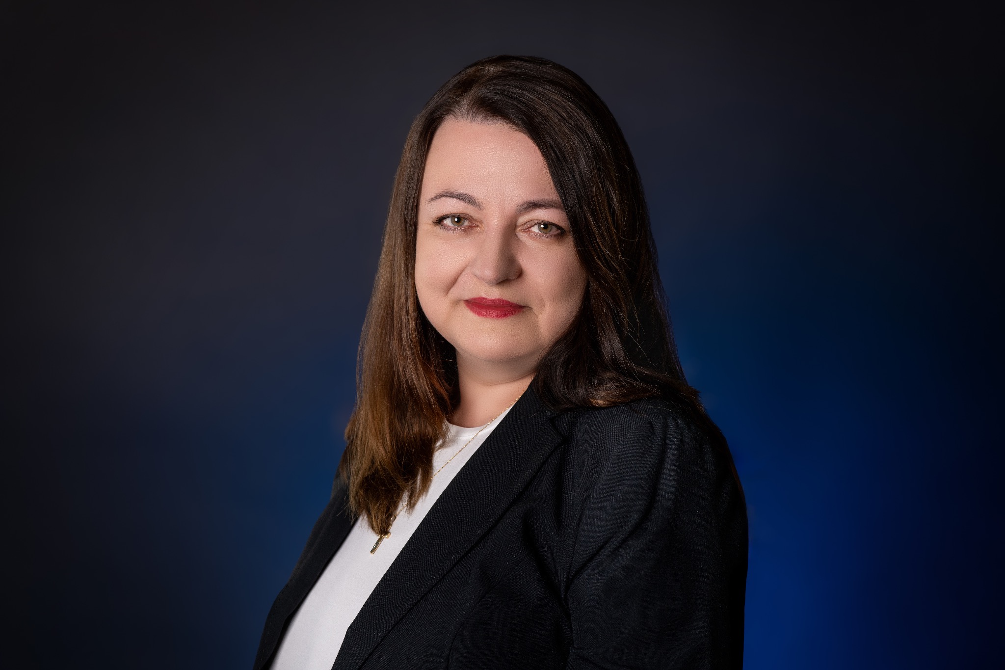 Portret kobiety w czarnej marynarce i białej bluzce, z delikatnym uśmiechem, na ciemnoniebieskim tle z subtelnym gradientem.