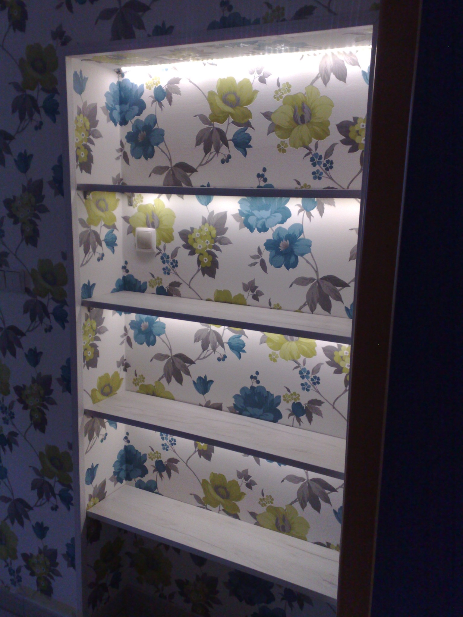 Wnęka z półkami, podświetlona taśmą LED, wykończona tapetą w kwiaty; widoczny fragment ściany z identyczną tapetą.