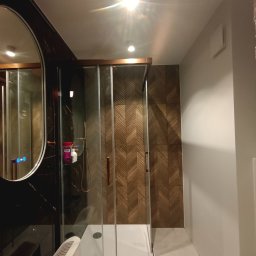 Krzysztof Karwowski - Prace Budowlane - Nowoczesna kabina prysznicowa z drewnopodobnymi płytkami w jodełkę, widoczna w łazience po remoncie w Szreńsku. Lustro z podświetleniem i marmurowa ściana.