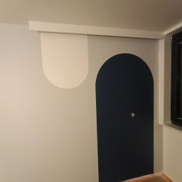 Krzysztof Karwowski - Prace Budowlane - Ściana po malowaniu z geometrycznymi wzorami: biały półokrąg i granatowy łuk. Widoczna listwa przypodłogowa i fragment okna. Minimalistyczny design wnętrza.