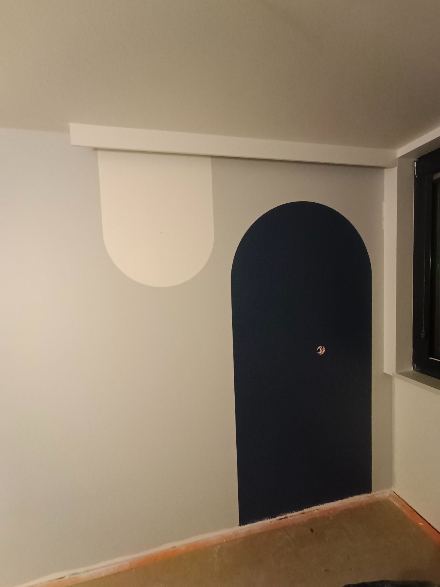 Ściana po malowaniu z geometrycznymi wzorami: biały półokrąg i granatowy łuk. Widoczna listwa przypodłogowa i fragment okna. Minimalistyczny design wnętrza.