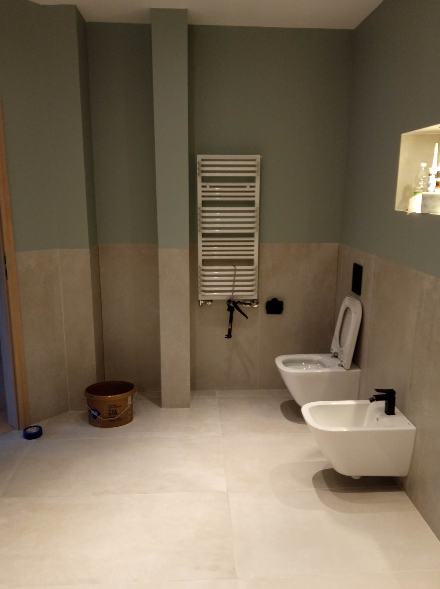 Jasna łazienka w trakcie wykańczania, widoczna toaleta, bidet z czarną armaturą, grzejnik drabinkowy, częściowo wykończone ściany z beżowymi płytkami i zieloną farbą, oraz wiadro z resztkami...