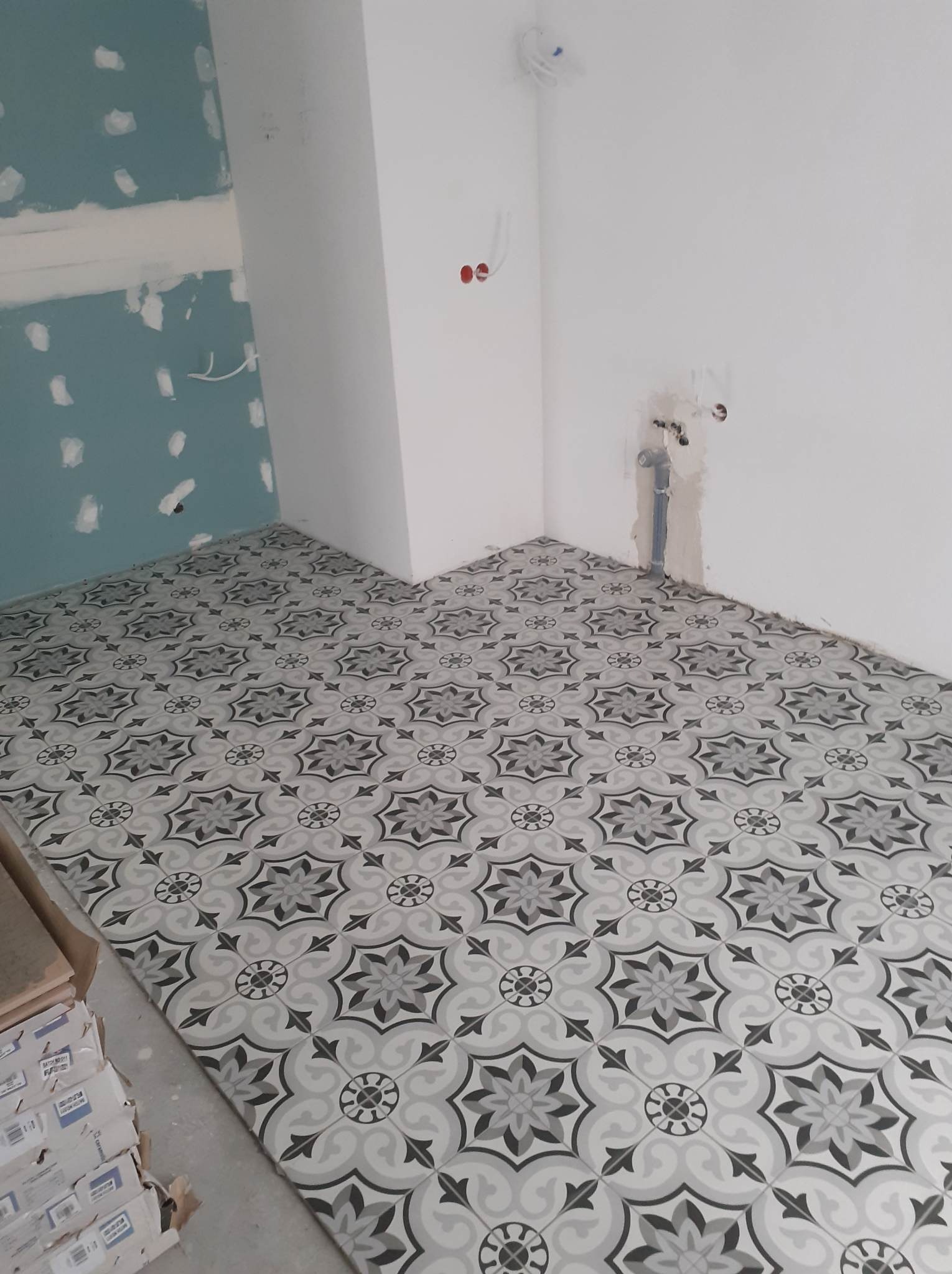 Ułożone płytki podłogowe w geometryczny wzór w nowo wykończonym pomieszczeniu, z widocznymi elementami instalacji elektrycznej i hydraulicznej w ścianach, oraz stosem opakowań płytek w rogu.