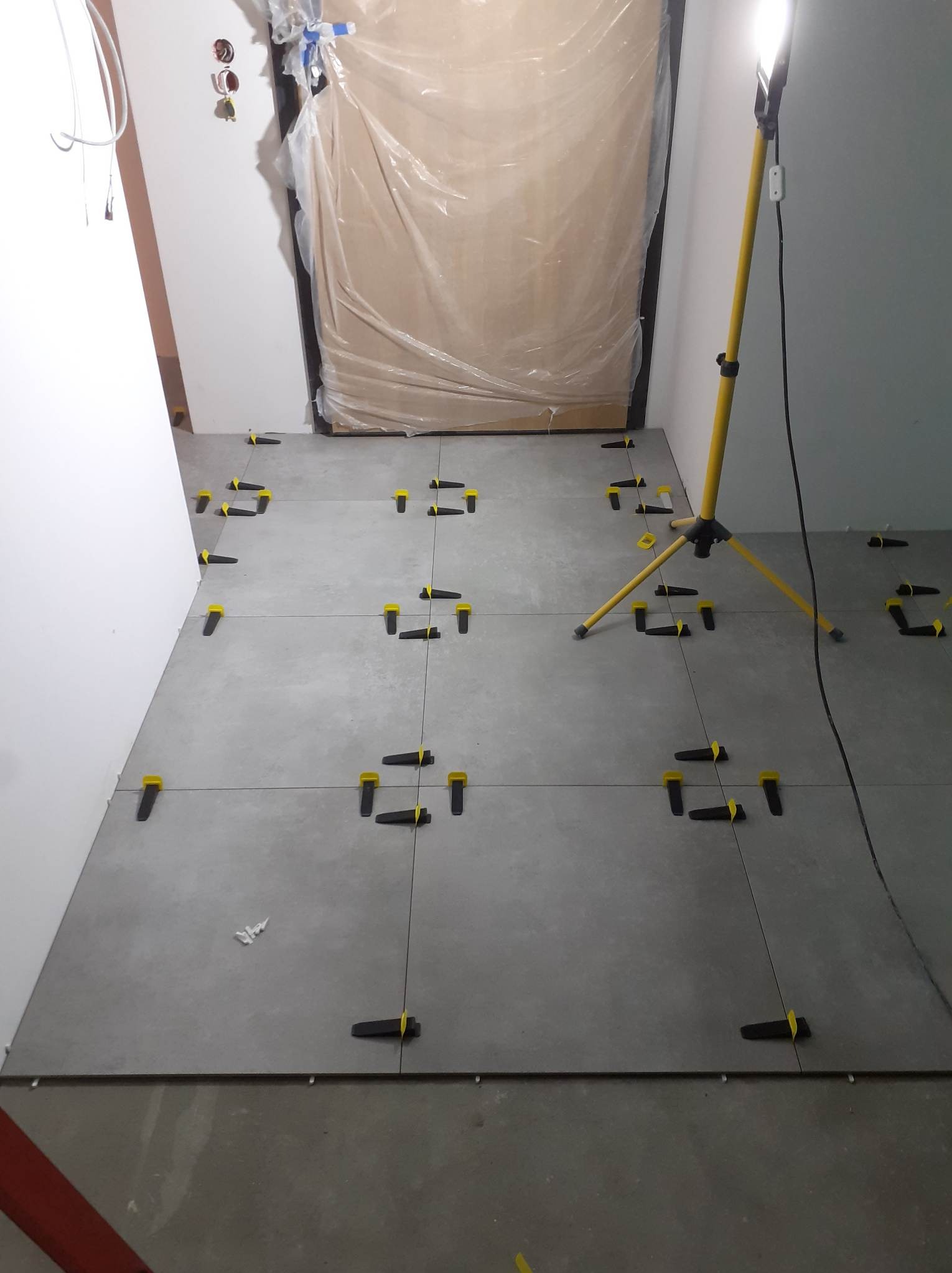 Szare płytki podłogowe w trakcie układania, z żółto-czarnymi klipsami poziomującymi, oświetlone lampą warsztatową, drzwi zabezpieczone folią malarską.