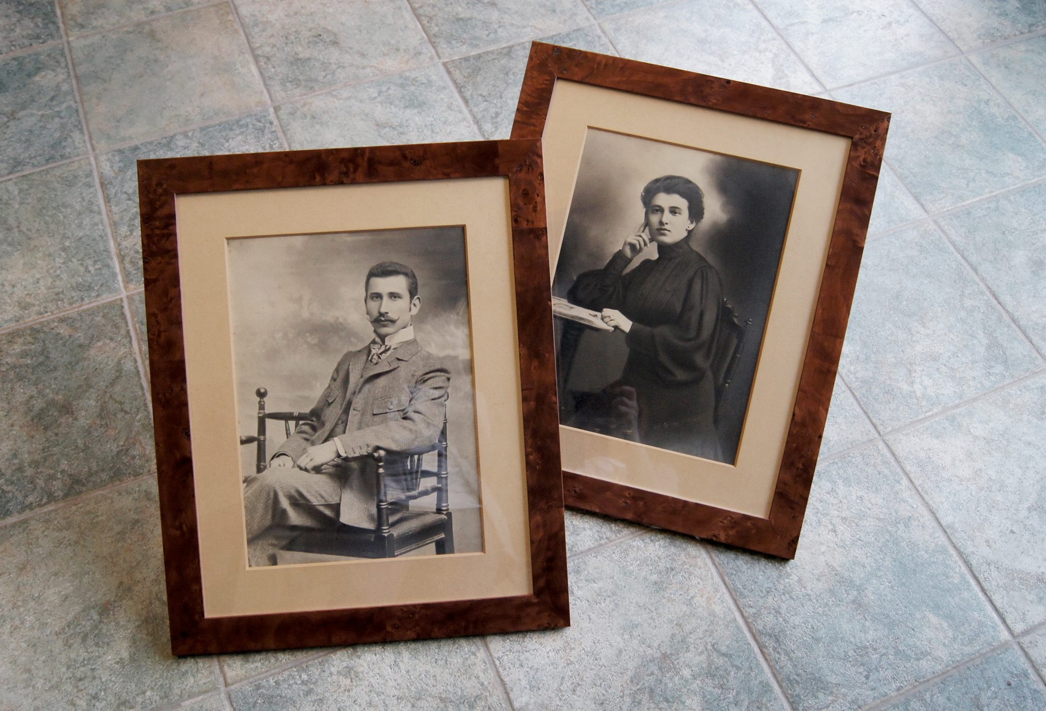 Dwa oprawione, czarno-białe portrety: mężczyzna z wąsami siedzący na krześle i kobieta opierająca się o stół, ułożone na podłodze z jasnych, kwadratowych płytek.