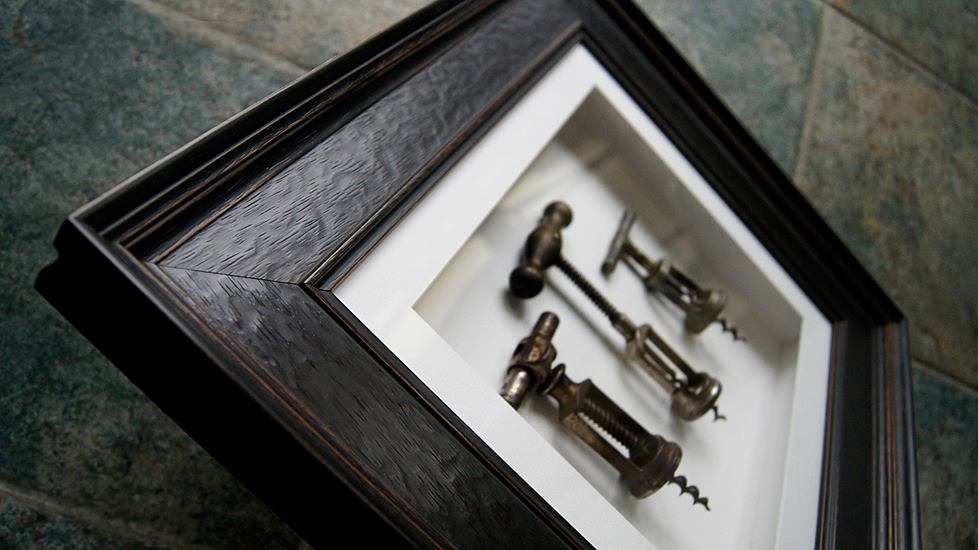 Dekoracja ścienna z trzema zabytkowymi korkociągami w ciemnej, drewnianej ramie, na tle ściany z kamiennych płytek.